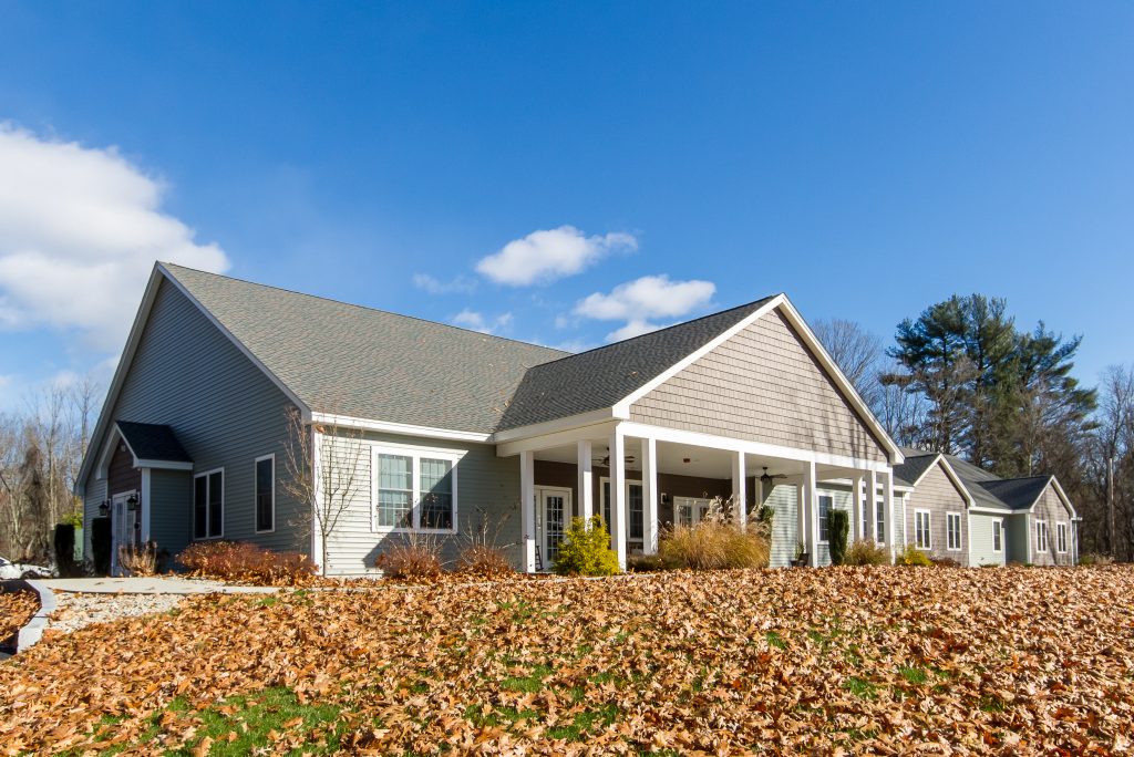 A tan building with white trim; in the foreground is a slight hill leading up to the house covered in orange autumn leaves, and in the background the sky is clear and blue.