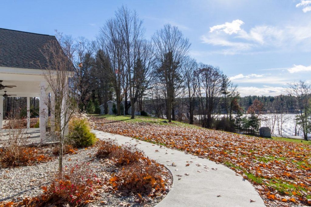 A walking path in autumn; to the far left is the entryway of a building, in the background on the right is a lake, and the path curves slightly to the left. There are red leaves all over the ground.