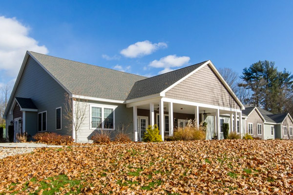 A tan building with white trim; in the foreground is a slight hill leading up to the house covered in orange autumn leaves, and in the background the sky is clear and blue.