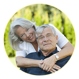 A white woman with silver hair is leaning over the sholder of a seated white man with grey hair; her arms are around his shoulders and her head is tilted to the side as they both smile at the camera.