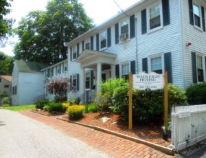 A white colonial style house with green shutters; the picture is taken from a sharp angle and shows the front of the house, the portico, and a brick walkway with landscaping.