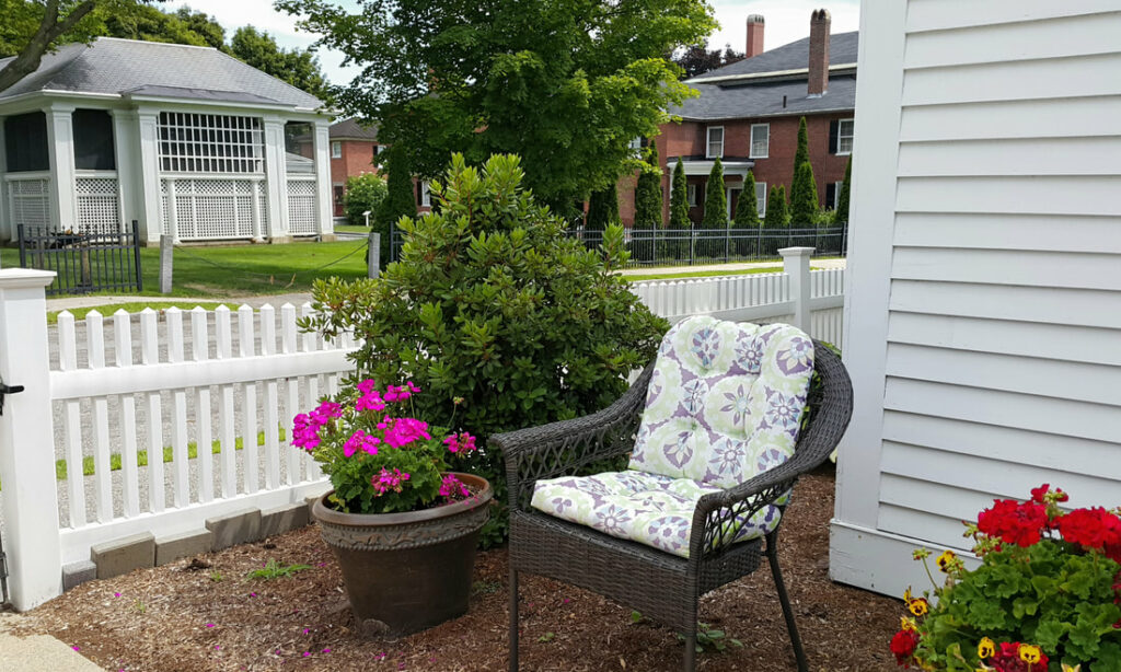 An outdoor seating area outside of a white building; there is a cushioned wicker chair on a landscaped ground, and to the left is a white picket fence