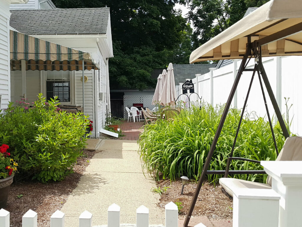 The view over a white fence into the private are between two buldings; on the left is a white building with green and tan awning, on the right is a tall white fence, and there is a brick path between them leading back to a patio area.