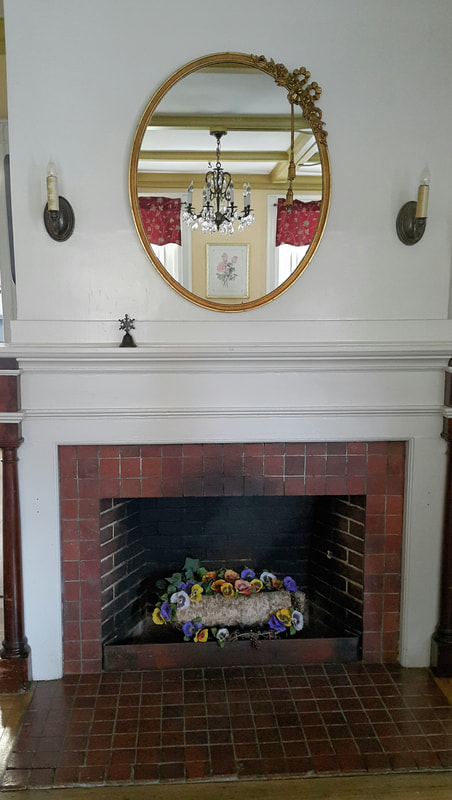 A brick fireplace with a simple white mantle; over the mantle is a large mirror showing the reflection of a chandelier, and in the hearth is a decorative log surrounded by flowers