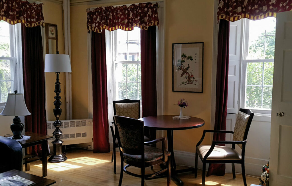 A sitting room painted with yellow walls; The windows are white and shuttered, and there are dark red curtains. To the left are two ornate lamps, and in the center of the image is a small table with three upholstered chairs