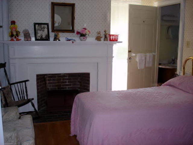 A small bedroom with a twin bed covered in a pink bedspread, a wooden rocking chair, a white armchair, and a large white brick fireplace; on the mantle is a photo and some knickknacks, and in the background to the right is an open door to a private bathroom.