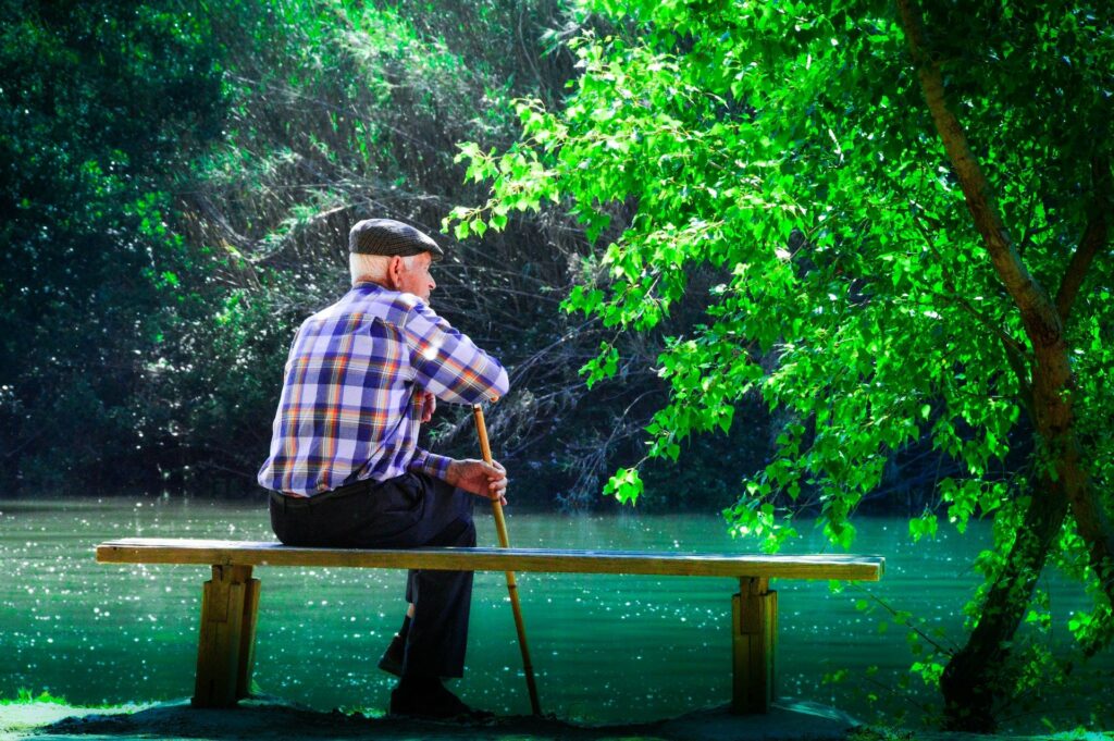 An elderly man wearing a plaid shirt and wool cap is sitting on a wooden bench overlooking a lake; he is leaning on a cane and looking off into the distance.