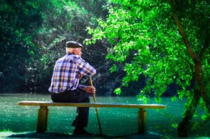 An elderly man wearing a plaid shirt and wool cap is sitting on a wooden bench overlooking a lake; he is leaning on a cane and looking off into the distance.