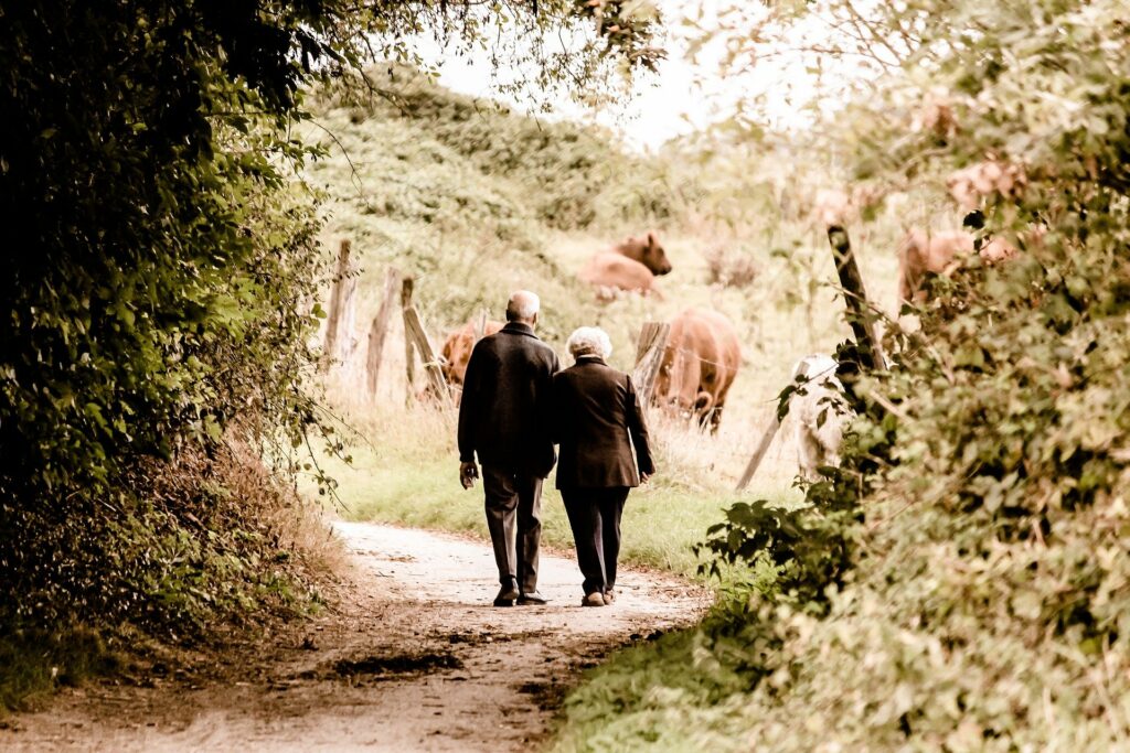 Two older people are walking hand in hand down a dirt path surrounded by bushes and trees.