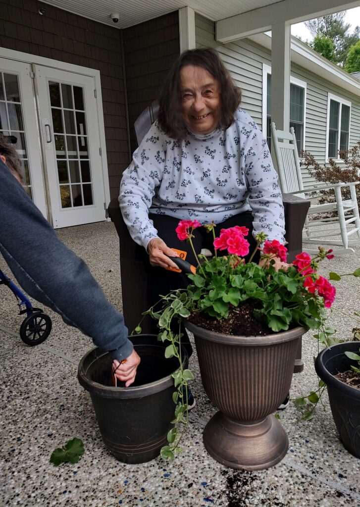 Woman sitting in a chair holding a shovel with a red potted geranium in front of her