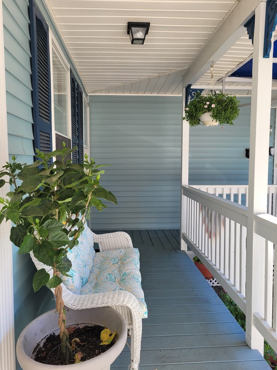 The covered porch area of Lincoln Terrace, showing the blue siding and white trim of the house, a cushoned wicker settee, and a large potted plant.