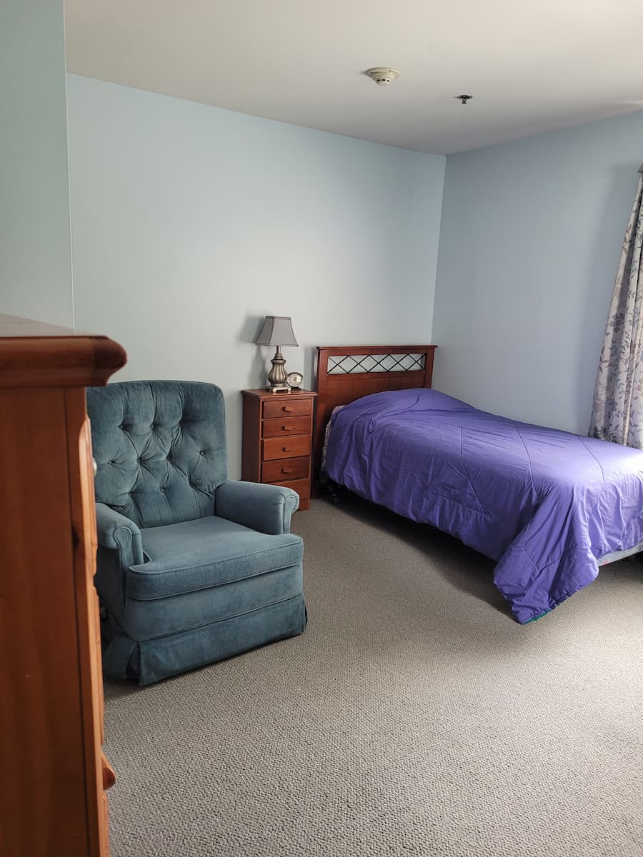 A bedroom with grey carpet and blue walls, with a grey armchair, a bed with dark blue coverlet, and warm wood furniture; at the right edge of the picture is the edge of blue floral curtains.