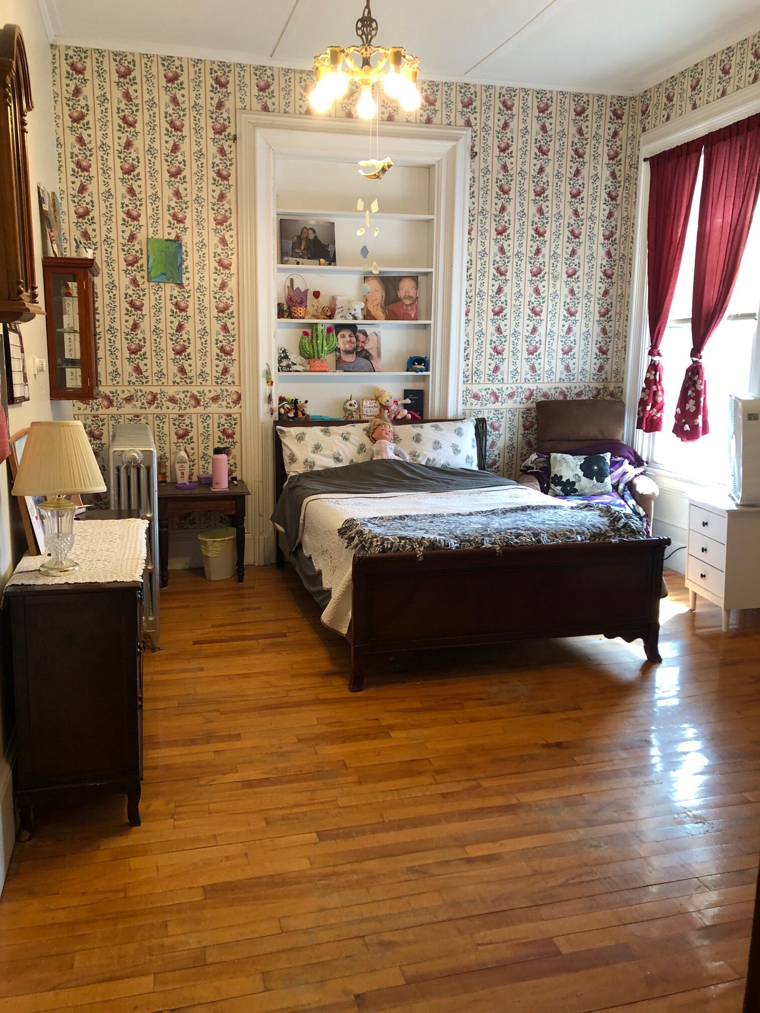 A bedroom decorated in colonial style, with hardwood floors, red and white floral wallpaper, red curtains, and dark brown wooden furniture; behind the bed is a bookshelf with photographs of family and friends