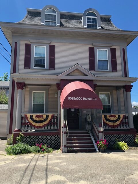 The front entry of a light brown house with grey and maroon trim; there is a maroon awning with white lettering reading "Rosewood Manor LLC"